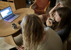 Trzy wychowanki siedzą na kanapie, patrzą na ekran laptopa, jedna z nich trzyma w dłoniach kubek z herbatą.