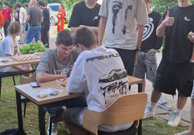 Dwóch chłopców siedzących, gra w szachy, kilkanaście innych stoi wokół nich lub obok służb  ratowników medycznych.