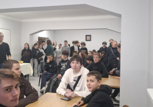 Kilkanaście osób siedzi na krzesłach, w oddali chłopcy stoją.