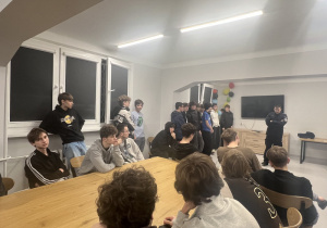 Chłopcy siedzą oraz stoją przy stolikach słuchając oraz zadając pytania.