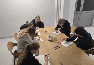 Przy stole siedzi grupa dziewcząt i chłopców, którzy rozwiązują zadania.