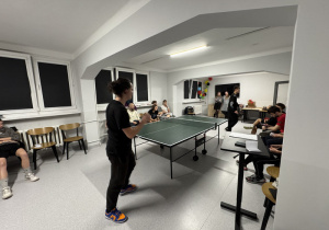 Dwóch wychowanków gra w tenisa stołowego, kilka osób siedząc obserwuje rozgrywkę