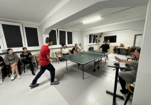 Dwóch wychowanków gra w tenisa stołowego, kilka osób siedząc obserwuje rozgrywkę