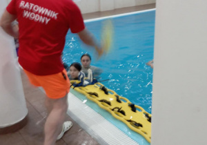 Ratowik stojący na brzegu basenu udziela instrukcji korzystania z deski ratowniczej