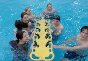 Cztery dziewczynki i czterech chłopców  ciągną w basenie deskę do ratowania życia
