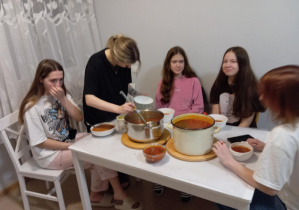 Jedna wychowanka nalewa zupę do talerzy. Cztery wychowanki siedzą przy stole i wspólnie jedzą ugotowany barszcz.