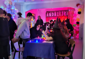 Kilkanaście osób stoi obserwuje wróżby,  ok 10 osób siedzi przy stole wróżąc sobie.