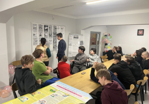 Wychowankowie siedzą przy stole a jeden z nich czyta informacje znajdujące się na tablicy dotyczące Praw Dziecka.