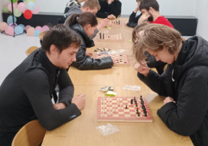 Młodzież siedzi przy stole na krzesłach i gra w szachy.