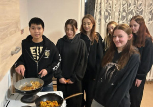 Chiński kucharz stoi przy piecu i uczy  wychowanki jak przygotować na woku kurczaka w pięciu smakach.