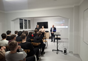 Wychowankowie siedzą na krzesłach i słuchają prelekcji.