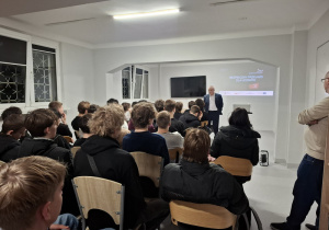 Wychowankowie siedzą na krzesłach i słuchają prelekcji.